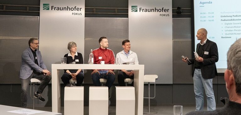 Vier Podiumsgäste und ein Redner auf einem Podium vor Bannern mit dem Logo von Fraunhofer Fokus
